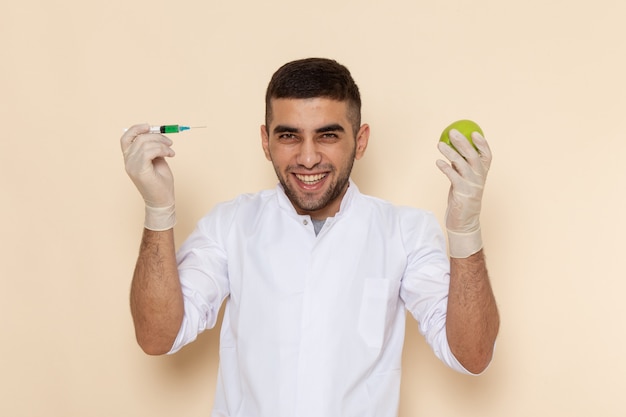 Front view young male in white suit wearing gloves injecting apple laughing on beige