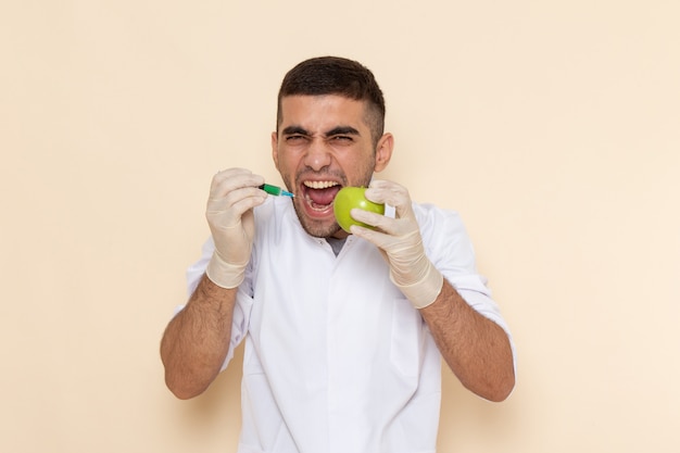 Front view young male in white suit wearing gloves injecting apple screaming on beige