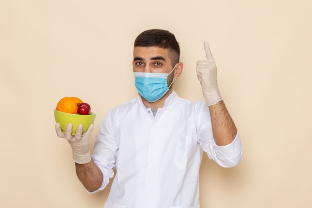 Front view young male in white suit wearing mask and gloves holding plate with fruits on beige