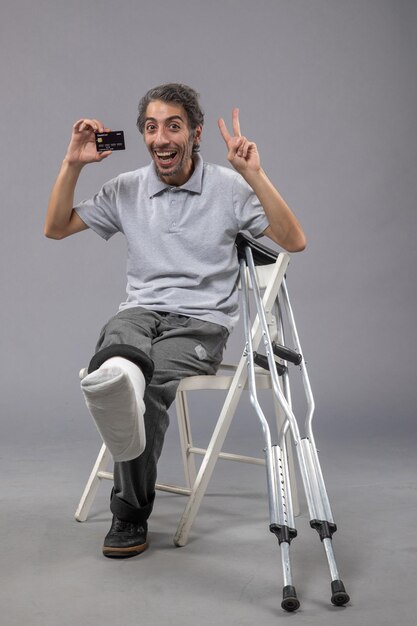 Front view young male with broken foot and bandage holding black bank card on grey wall human twist male accident foot pain