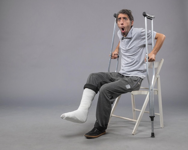 Front view young male with broken foot trying to stand up holding crutches on a grey wall foot broken pain twist accident leg