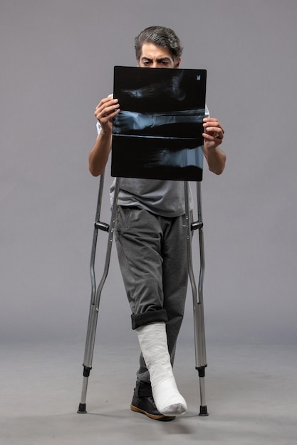 Front view young male with broken foot using crutches and holding his x-ray on grey wall disable broken accident pain foot twist