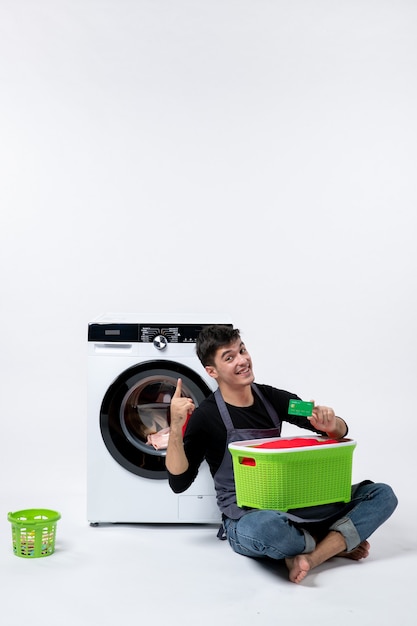 Front view of young male with green basket full of clothes holding bank card on the white wall