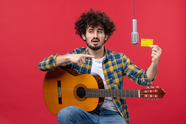 Front view young male with guitar holding bank card on red wall live performance musician concert band money