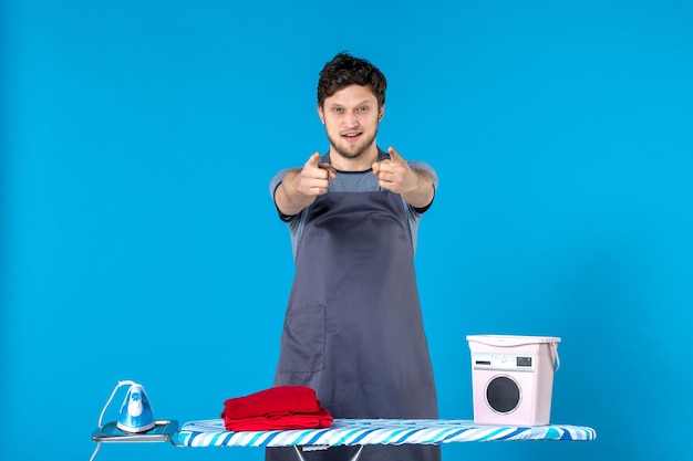 Free photo front view young male with ironing board and clothes pointing on blue background color housework laundry cleaning washing machine