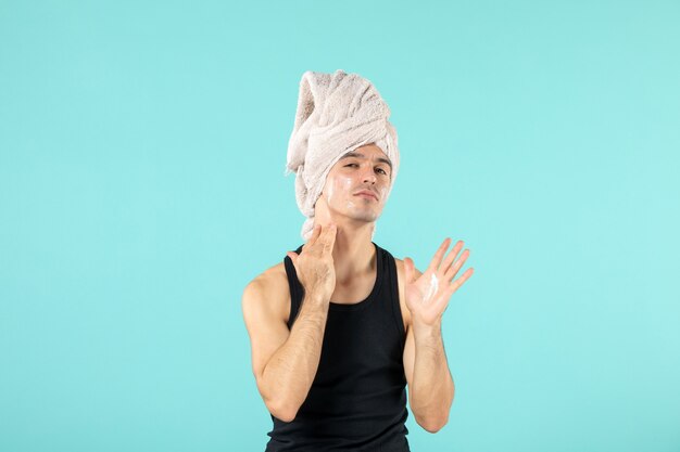 front view of young man after shower applying cream to his face on blue wall