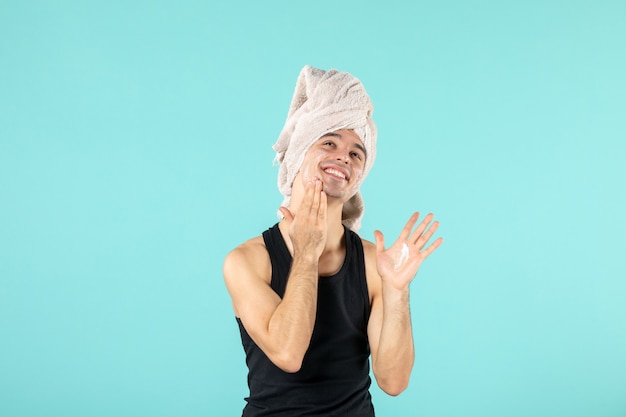 front view of young man after shower applying cream to his face on blue wall