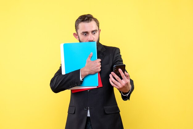 Front view of young man in black suit holding folders and looking scared at a phone on yellow