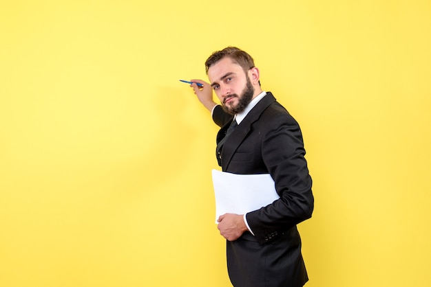 Front view of young man in black suit pointing to the yellow wall using a pen