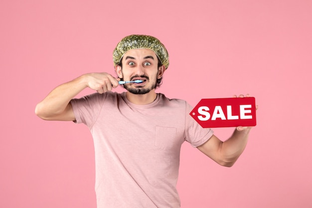 front view of young man cleaning his teeth and holding sale banner on a pink wall