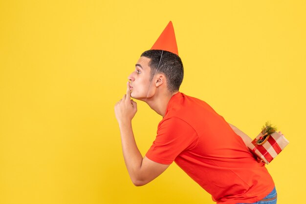 Front view of young man hiding xmas present behind his back on yellow wall