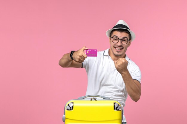 Front view of young man holding bank card on summer vacation on pink wall