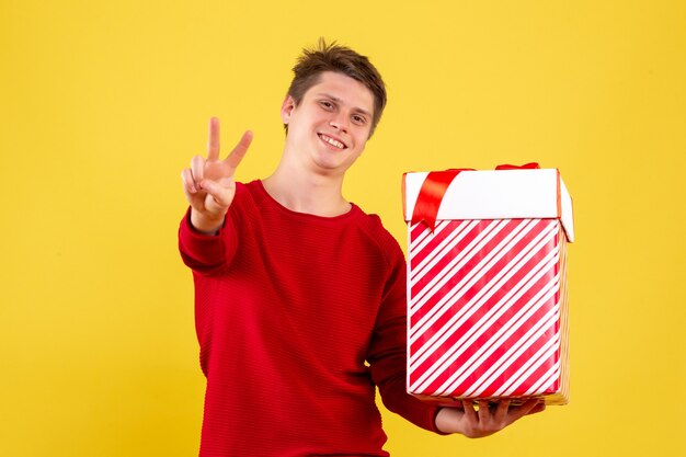 Front view of young man holding big xmas present smiling on yellow wall