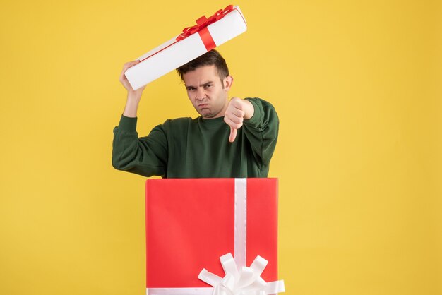 Front view young man holding box cover making thum down sign standing behind big giftbox on yellow 