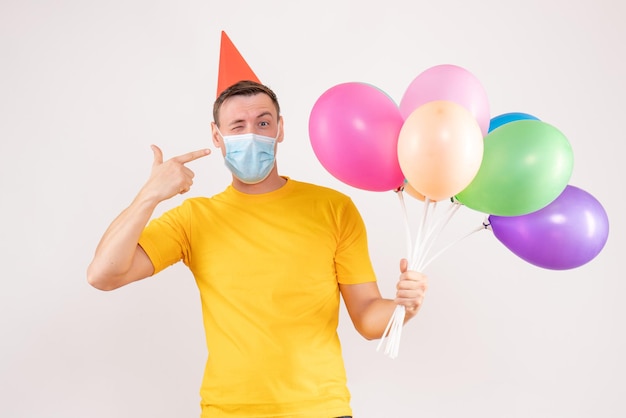 Front view of young man holding colorful balloons in mask on a white wall