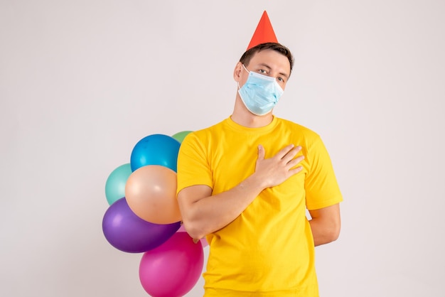 Front view of young man holding colorful balloons in mask on white wall