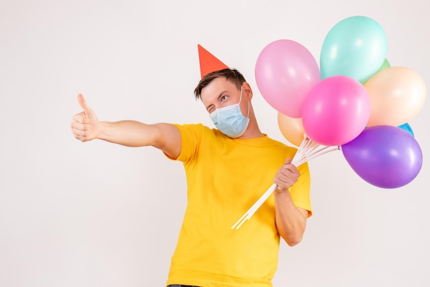 Front view of young man holding colorful balloons in mask on white wall