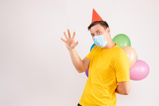 Front view of young man holding colorful balloons in mask on white wall