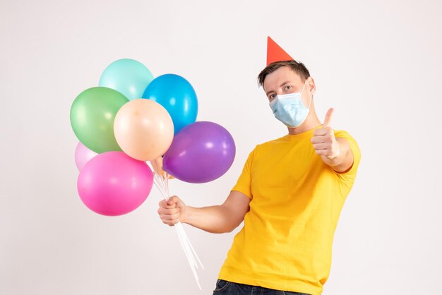 Front view of young man holding colorful balloons in sterile mask on white wall