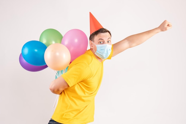 Front view of young man holding colorful balloons in sterile mask on white wall