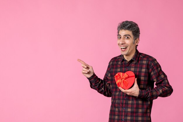 Front view young man holding red heart shaped present on pink wall