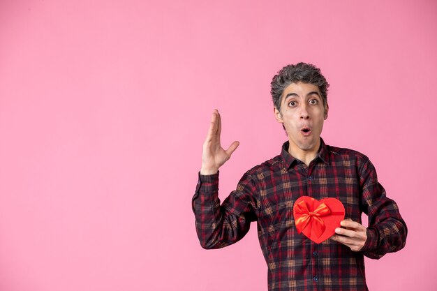 Front view young man holding red heart shaped present on pink wall