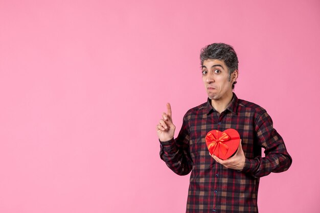 Front view young man holding red heart shaped present on pink wall