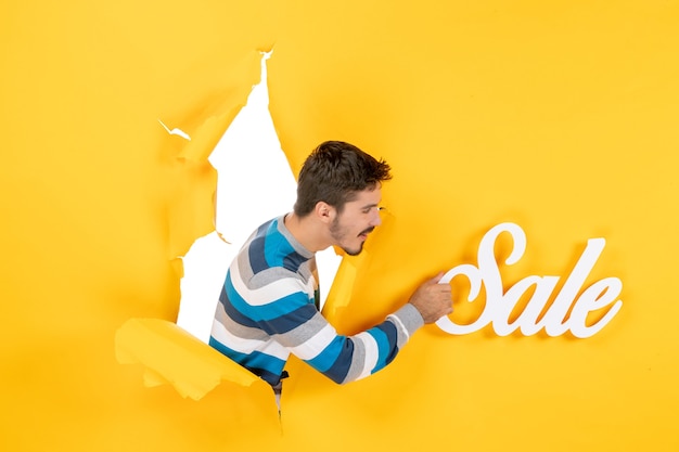 Free Photo front view young man holding sale letters through the round shaped hole in the yellow wall