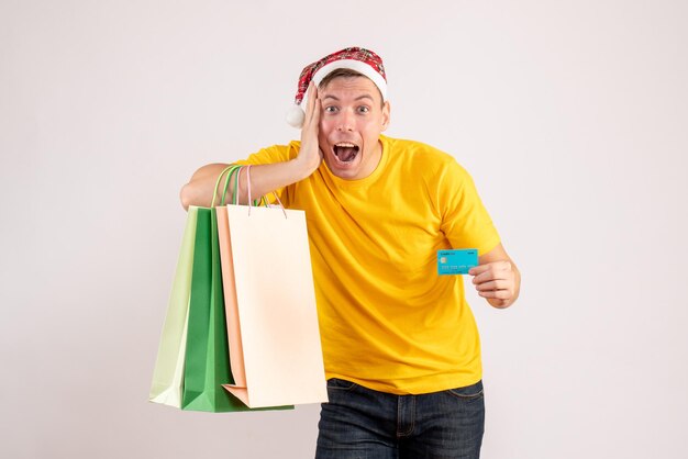 Front view of young man holding shopping packages and bank card on white wall