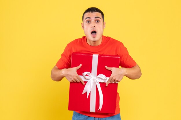 Front view of young man holding xmas present with surprised expression on yellow wall