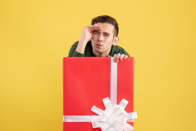 Front view young man looking at something standing behind big giftbox on yellow 