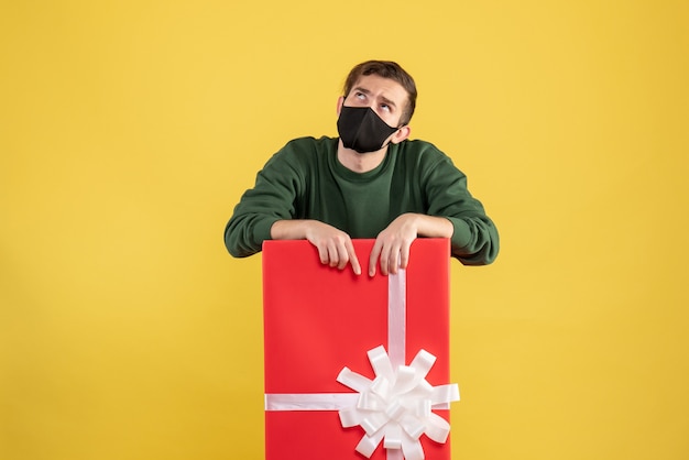 Front view young man lookng up standing behind big giftbox on yellow 