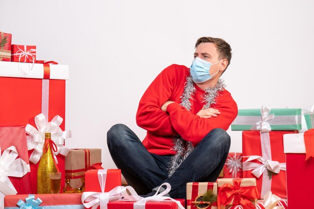 Front view of young man in mask sitting around xmas presents on a white wall