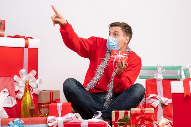 Front view of young man in mask sitting around xmas presents on a white wall