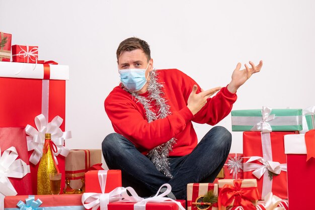 Front view of young man in mask sitting around xmas presents on the white wall