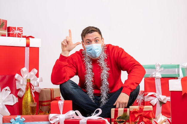 Front view of young man in mask sitting around xmas presents on white wall