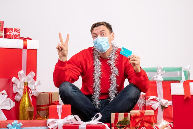 Front view of young man in mask sitting around xmas presents on white wall