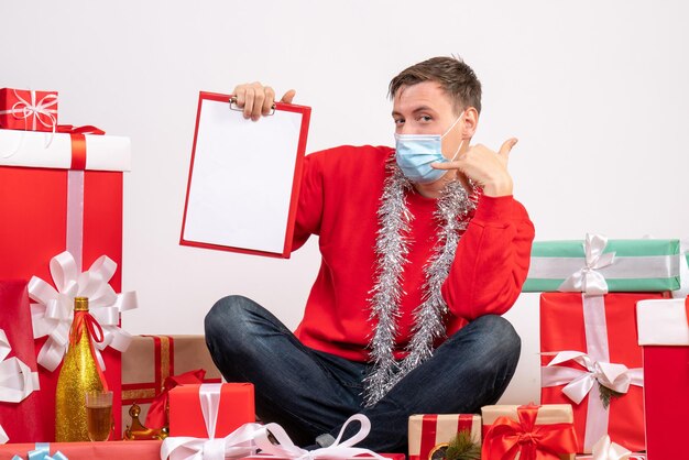 Front view of young man in mask sitting around xmas presents with note on a white wall