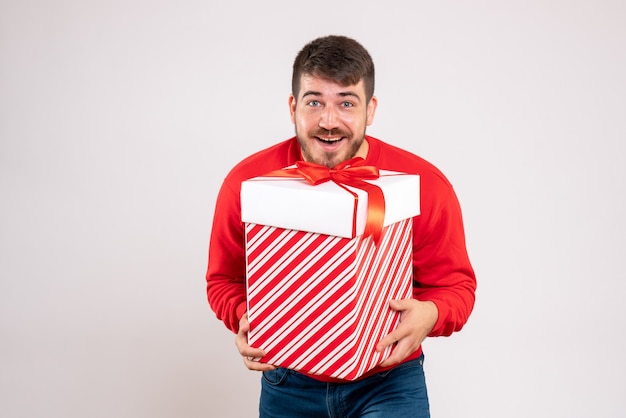 Front view of young man in red shirt holding christmas present in box on white wall