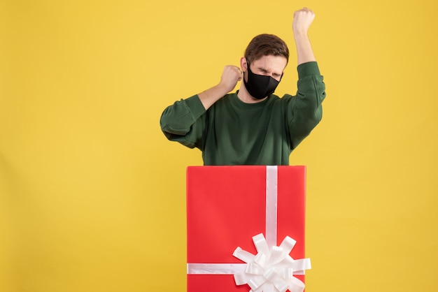 Free photo front view young man showing winning gesture standing behind big giftbox on yellow