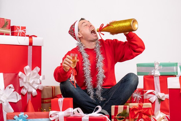 Front view of young man sitting around presents drinking champagne on white wall