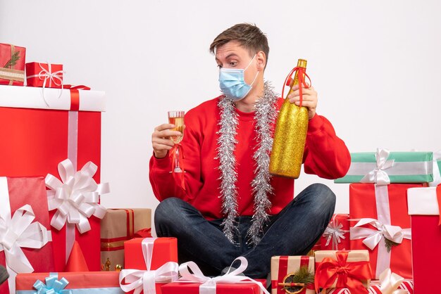 Front view of young man sitting around xmas presents celebrating with champagne on white wall