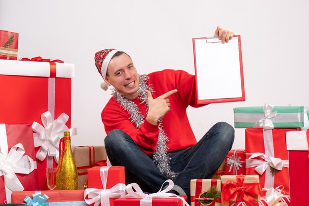 Front view of young man sitting around xmas presents with note on the white wall