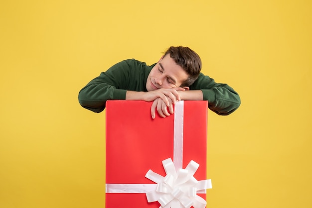 Front view young man sleeping behind big giftbox putting head on box on yellow 