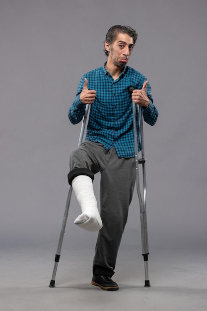 Free photo front view young man using crutches due to broken foot on grey desk accident disabled broke damage leg