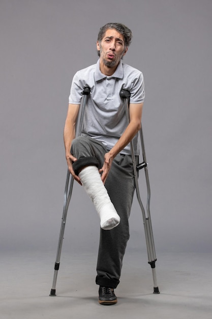 Front view young man using crutches due to broken foot on grey desk disable leg foot broken accident