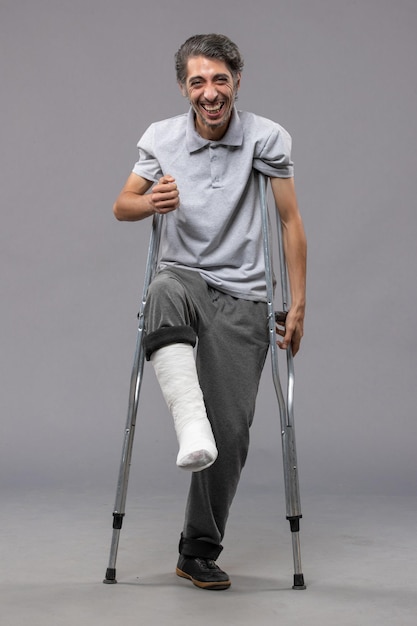 Front view young man using crutches due to broken foot on grey wall accident disable broke leg broken