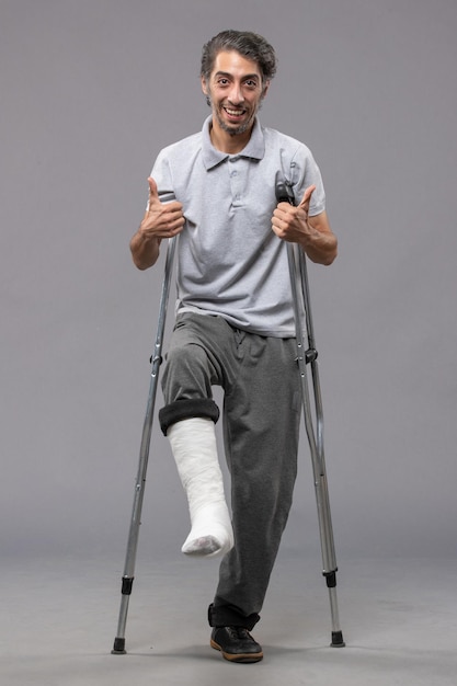 Free photo front view young man using crutches due to broken foot on grey wall accident disable broke legs broken