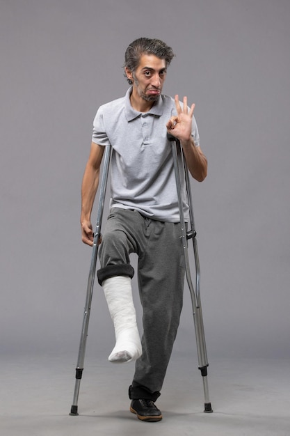 Front view young man using crutches due to broken foot on grey wall disable broke leg broken accident