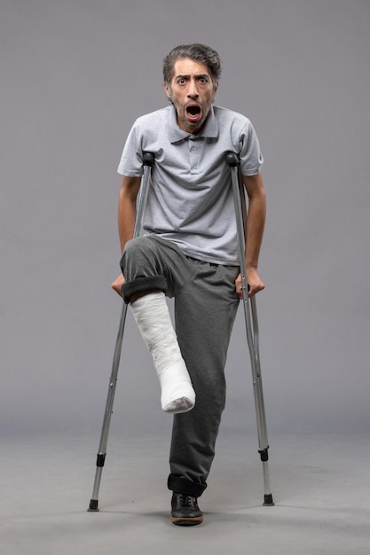 Free photo front view young man using crutches due to broken foot on a grey wall disable leg foot broken accident